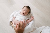 Сверху обрезанная неузнаваемая рука, держащая очаровательного новорожденного спящего, лежащего на мягком одеяле в корзине, положенной на пол — стоковое фото
