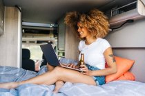 Молодая афроамериканка-путешественница с вьющимися волосами пьет пиво и смотрит кино на ноутбуке во время летних каникул в кемпере — стоковое фото