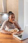 Afro-americana mujer disfrutando sabroso patacon con cobertura mientras navega por Internet en netbook en la cocina en casa - foto de stock