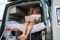 Jeune voyageuse afro-américaine avec des cheveux bouclés boire de la bière et regarder un film sur ordinateur portable tout en se reposant à l'intérieur du camping-car pendant les vacances d'été — Photo de stock