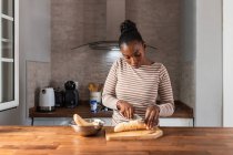Femme noire en vêtements avec ornement rayé coupant banane de cuisson avec couteau sur planche à découper à table dans la maison — Photo de stock