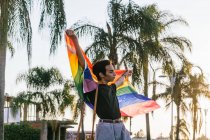 Content gay male walking with rainbow LGBT flag in raised hands along street in tropical city — Stock Photo