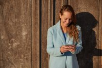 Happy adult woman in blue coat leaning on old building while browsing on cellphone in city district in sunny day — Stock Photo