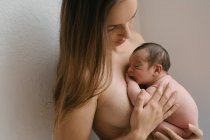 Vista lateral de la tierna madre en topless con los ojos cerrados de pie con lindo bebé desnudo cerca de la pared en casa - foto de stock