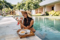 Cheerful female tourist in swimwear enjoying refreshing drink while leaning on poolside with tasty breakfast on tray in tropical resort — Stock Photo
