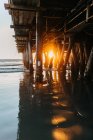 Brillante luz del sol en la noche penetrando pilas de muelle de Santa Mónica con olas pacíficas del océano que corren en la playa en California - foto de stock