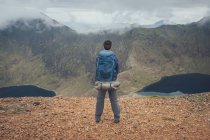 Vista posterior del viajero masculino anónimo con la mochila parada en el mirador en las tierras altas y admirando paisajes increíbles de la cordillera durante el trekking en Gales en el día nublado - foto de stock