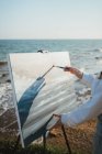 Cortar jovem mulher de pé na costa gramada perto de areia e oceano em dia ensolarado enquanto desenha imagem com escova na tela no cavalete — Fotografia de Stock