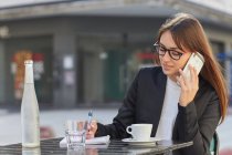 Jovem empresária positiva em terno elegante e óculos tomando notas no caderno durante a conversa telefônica enquanto se senta à mesa no café ao ar livre na cidade — Fotografia de Stock