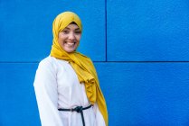 Vue latérale de la femme musulmane branchée en hijab jaune debout regardant la caméra dans la rue sur fond bleu — Photo de stock