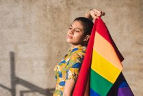 Mujer étnica bisexual joven seria con bandera multicolor que representa símbolos LGBTQ y mira hacia abajo en el día soleado - foto de stock