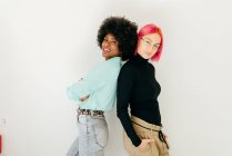 Веселая молодая розововолосая женщина и афро-американская девушка в стильном наряде, стоящие спиной к спине на белом фоне — стоковое фото