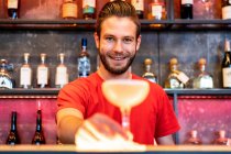 Barman alegre servir vidrio con cóctel de alcohol en el mostrador en el bar y mirando a la cámara - foto de stock