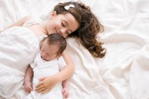 Вид зверху весела маленька дівчинка, що обіймає чарівного немовляти, лежачи на м'якому ліжку вдома — стокове фото