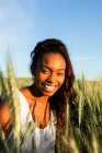 Молода чорна леді в білій літній сукні, що йде на зелене пшеничне поле, дивлячись на камеру вдень під блакитним небом — стокове фото