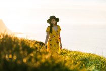 Attraktive junge Frau in gelbem Kleid und Hut steht auf einer grünen Wiese in sonniger Landschaft — Stockfoto