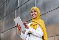 Smiling Muslim female entrepreneur in yellow hijab standing in street and browsing tablet — Stock Photo