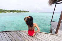 Rückansicht einer nicht wiedererkennbaren Frau im Badeanzug, die auf einem Holzsteg sitzt und sich auf den Malediven entspannt — Stockfoto