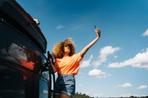 De baixo mulher negra com cabelo encaracolado em pé na escada fora van e tomando selfie contra céu azul nublado durante a viagem de carro — Fotografia de Stock