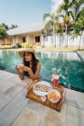 Cheerful female tourist leaning on poolside while drinking coffee against tray with yummy breakfast in sunlight — Stock Photo