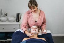 Cosmetologist applying moisturizing facial mask on face of female customer during skincare procedure in modern beauty salon — Stock Photo