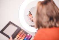Избыточный вес женщины с палитрой нанесения красочных пигментов на лицо при взгляде на зеркало рядом с кольцом света в студии — стоковое фото