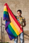 Mujer étnica bisexual joven seria con bandera multicolor que representa símbolos LGBTQ y mira a la cámara en un día soleado - foto de stock