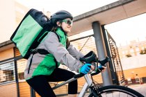 Низький кут жіночого кур'єра з термальною сумкою, посміхаючись і їздячи на велосипеді на вулиці, роблячи доставку в сонячний день у місті — стокове фото