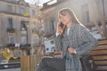 Femme millénaire moderne en tenue de printemps élégant assis sur le banc et répondre à un appel téléphonique tout en se reposant sur la rue urbaine regardant loin — Photo de stock