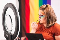Más tamaño femenino mirando el espejo con luz de anillo y aplicando maquillaje creativo contra la bandera LGBT en el estudio - foto de stock