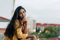 Молодая латиноамериканка в повседневной одежде опирается на перила и смотрит в камеру, расслабляясь вечером на балконе в городе — стоковое фото