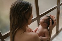 Vista lateral de la tierna madre con los ojos cerrados de pie con lindo bebé desnudo cerca de la ventana en casa - foto de stock
