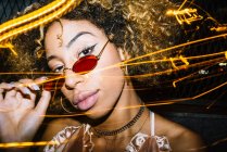 Autosicuro giovane donna afroamericana con i capelli ricci in occhiali da sole alla moda e top rilassante in strada in serata vicino a congelare le luci — Foto stock