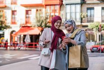 Muslim female friends in hijabs and with paper bags using smartphone on street after shopping — Stock Photo