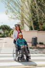 Взрослая женщина толкает инвалидное кресло со старшей матерью и пересекает дорогу в городе во время прогулки летом — стоковое фото