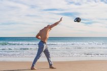 Vue latérale de l'homme sportif anonyme sans chemise jetant kettlebell sur la côte sablonneuse avec des vagues de l'océan sur le fond — Photo de stock