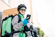 Молодая веселая женщина с термальной сумкой просматривает смартфон с GPS-картой, сидя на велосипеде для доставки пищи на городской улице — стоковое фото