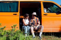Encantada pareja de viajeros sentados en furgoneta y mirando a través de fotos en la cámara durante la aventura de verano - foto de stock