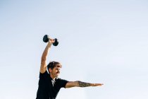 Atleta masculino concentrado em sportswear trabalhando com halteres enquanto olha para a frente sob o céu claro — Fotografia de Stock
