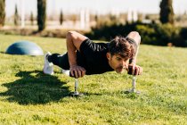 Взрослый спортсмен тренируется на гантели, стоя в позе доски и глядя вперед на городской газон под солнечным светом — стоковое фото