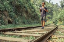 Cheerful male traveler with backpack walking along railway in tropical woods during summer vacation — Stock Photo