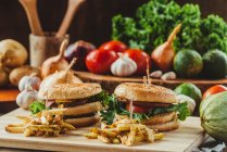 Hambúrgueres apetitosos com legumes e costeletas colocados em tábua de madeira com batatas fritas na cozinha — Fotografia de Stock