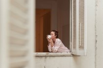 Tranquillo femminile in pigiama appoggiato alla finestra con tazza di caffè del mattino e occhi chiusi — Foto stock