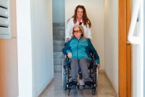 Mujer adulta satisfecha empujando silla de ruedas con madre mayor y caminando por el pasillo en el centro de rehabilitación - foto de stock