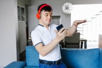 Entzückter lateinischer Teenie-Junge mit Kopfhörer auf Videoanruf auf Handy, während er in der Nähe des Sofas zu Hause steht und in die Kamera schaut — Stockfoto