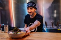 Bearded male in bandana and black t shirt putting plate with noodle on wooden counter for client in ramen bar — Stock Photo