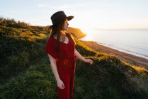 Attraente giovane femmina in abito da sole rosso e cappello in piedi su verdeggiante prato erboso in campagna soleggiata — Foto stock