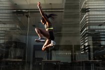 Вид збоку сильної чорної жінки, що стрибає високо біля скляної стіни сучасної будівлі під час тренувань на вулиці міста в сонячний день — стокове фото