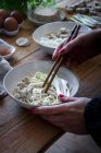 Cultivé personne méconnaissable préparant des nouilles ramen fraîches cuites avec du tofu, des œufs et des légumes avec des baguettes sur une table en bois — Photo de stock