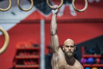 Starker, hemdsloser Mann, der während des intensiven Trainings in einem modernen Fitnessstudio auf Gymnastikringen stehend in die Kamera blickt — Stockfoto